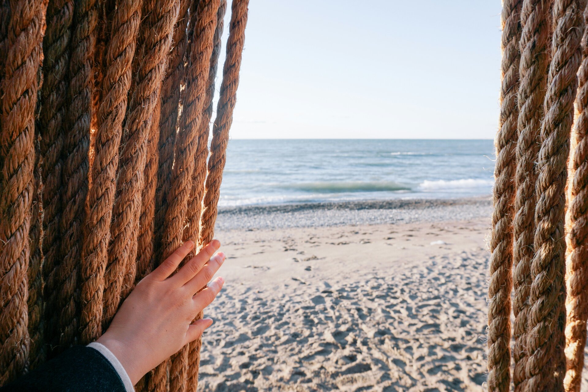 Man blickt auf einen Strand und das Meer, im Vordergrund schiebt eine Hand einen Vorhang zur Seite